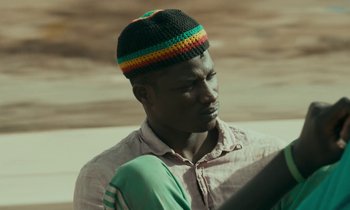 Movie still from “Atlantics” (2019), directed by Mati Diop – A man wearing a colorful hat and a green shirt; Close Up shot, Low angle