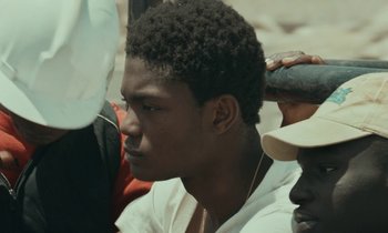 Movie still from “Atlantics” (2019), directed by Mati Diop – A young man with a black afro is sitting in a crowd; Close Up shot, Over the shoulder angle