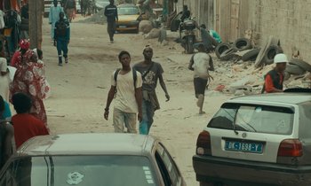 Movie still from “Atlantics” (2019), directed by Mati Diop – A group of people walking down a street next to a car; Wide shot, Over the shoulder angle