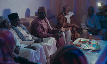 Movie still from “Atlantics” (2019), directed by Mati Diop – A group of men sitting on top of couches in a living room; Medium shot, Over the shoulder angle