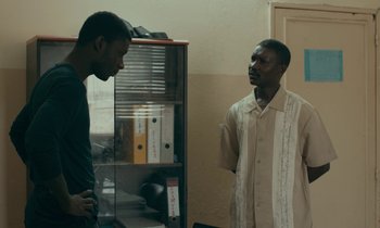 Movie still from “Atlantics” (2019), directed by Mati Diop – Two men standing next to each other in front of a book case; Medium shot, Over the shoulder angle