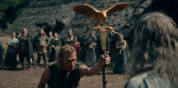 Movie still from “Barbarians” (2020), created by Arne Nolting – A man holding a wooden stick with an owl on it; Medium shot, Low angle