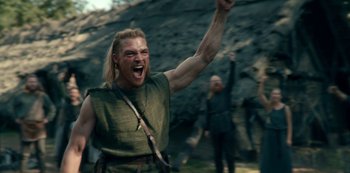 Movie still from “Barbarians” (2020), created by Arne Nolting – A man with a long blonde hair and a green shirt is raising his fist in the air; Medium shot, Low angle