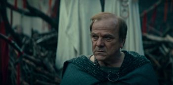 Movie still from “Barbarians” (2020), created by Arne Nolting – An older man wearing a green cape and a chain around his neck; Close Up shot, Over the shoulder angle