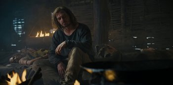 Movie still from “Barbarians” (2020), created by Arne Nolting – A man sitting on a couch next to an open fire; Medium shot, Low angle