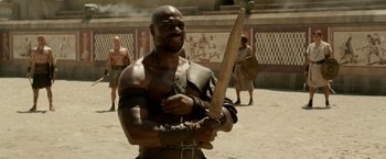 Movie still from “Pompeii” (2014), directed by Paul W.S. Anderson – A man holding a wooden sword in front of other men; Medium shot, Over the shoulder angle