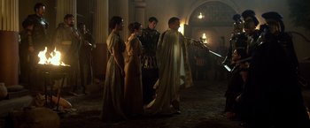 Movie still from “Pompeii” (2014), directed by Paul W.S. Anderson – A group of people standing in a room with a sword in their hand; Wide shot, Over the shoulder angle