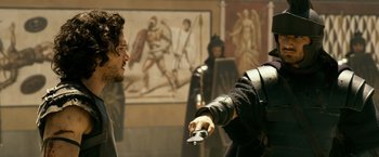 Movie still from “Pompeii” (2014), directed by Paul W.S. Anderson – Two men are holding a remote control in front of a painting; Medium shot, Over the shoulder angle