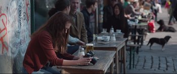 Movie still from “Berlin Syndrome” (2017), directed by Cate Shortland – A woman sitting at a wooden table with a camera; Medium shot, Over the shoulder angle