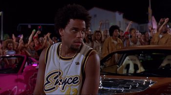 Movie still from “2 Fast 2 Furious” (2003), directed by John Singleton – A man in a basketball jersey sitting in front of a crowd of onlookers; Medium shot, Over the shoulder angle