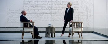 Movie still from “6 Underground” (2019), directed by Michael Bay – A woman standing in front of a man sitting in a chair; Wide shot, Low angle