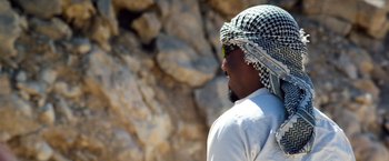 Movie still from “6 Underground” (2019), directed by Michael Bay – A man wearing a keffiyeh is looking at a rock wall; Close Up shot, Over the shoulder angle