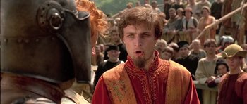 Movie still from “A Knight's Tale” (2001), directed by Brian Helgeland – A man with a beard and a red jacket is standing in front of a crowd of people; Close Up shot, Over the shoulder angle