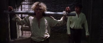 Movie still from “A Knight's Tale” (2001), directed by Brian Helgeland – A man chained up to a wooden pole; Medium shot, Over the shoulder angle