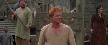Movie still from “A Knight's Tale” (2001), directed by Brian Helgeland – A man with red hair is standing in front of a wooden wall; Medium shot, Low angle