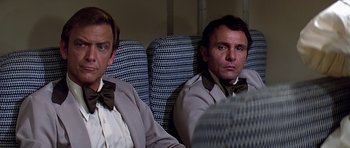 Movie still from “Airport '77” (1977), directed by Jerry Jameson – A couple of men sitting next to each other on some seats; Medium shot, Over the shoulder angle