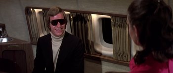 Movie still from “Airport '77” (1977), directed by Jerry Jameson – A man in a suit and sunglasses standing in front of a window; Medium shot, Low angle