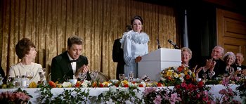 Movie still from “Airport” (1970), directed by George Seaton – A woman standing at a table in front of a microphone; Medium shot, Over the shoulder angle