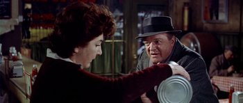 Movie still from “Airport” (1970), directed by George Seaton – An older man in a hat is holding a plate while an older woman in a sweater looks on; Medium shot, Over the shoulder angle