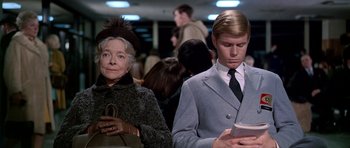 Movie still from “Airport” (1970), directed by George Seaton – A man and a woman looking at a cell phone in front of a crowd; Medium shot, Low angle