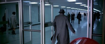 Movie still from “Airport” (1970), directed by George Seaton – A man in a long coat and a hat walking through an airport; Wide shot, Over the shoulder angle