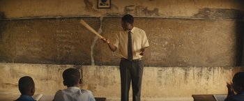 Movie still from “Beasts of No Nation” (2015), directed by Cary Joji Fukunaga – A man holding a baseball bat in front of a chalkboard; Medium shot, Low angle