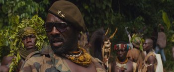Movie still from “Beasts of No Nation” (2015), directed by Cary Joji Fukunaga – A man wearing a hat and sunglasses with other people in the background; Close Up shot, Low angle