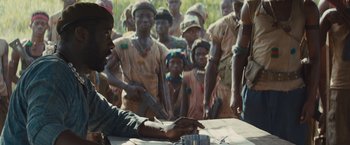Movie still from “Beasts of No Nation” (2015), directed by Cary Joji Fukunaga – A group of people standing around a table; Medium shot, Over the shoulder angle