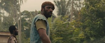 Movie still from “Beasts of No Nation” (2015), directed by Cary Joji Fukunaga – A man with a beard and a hat is standing in the woods; Medium shot, Low angle