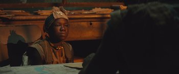 Movie still from “Beasts of No Nation” (2015), directed by Cary Joji Fukunaga – A young boy sitting at a table in front of an old desk; Close Up shot, Over the shoulder angle