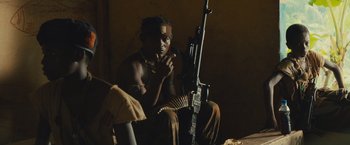Movie still from “Beasts of No Nation” (2015), directed by Cary Joji Fukunaga – A man sitting in front of a rifle; Medium shot, Over the shoulder angle