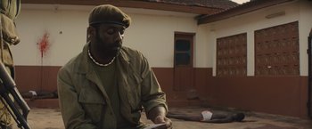 Movie still from “Beasts of No Nation” (2015), directed by Cary Joji Fukunaga – A man sitting on the ground looking down at a piece of paper; Medium shot, High angle