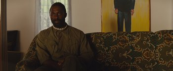 Movie still from “Beasts of No Nation” (2015), directed by Cary Joji Fukunaga – A man sitting on top of a couch in a living room; Medium shot, Over the shoulder angle