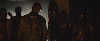 Movie still from “Beasts of No Nation” (2015), directed by Cary Joji Fukunaga – A man standing next to another man in a dark room; Medium shot, Low angle