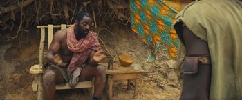 Movie still from “Beasts of No Nation” (2015), directed by Cary Joji Fukunaga – A man sitting on top of a wooden bench next to a bowl; Medium shot, High angle
