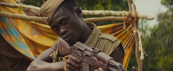 Movie still from “Beasts of No Nation” (2015), directed by Cary Joji Fukunaga – A man holding an ak - 4 7 rifle while standing in a hammock; Close Up shot, Low angle