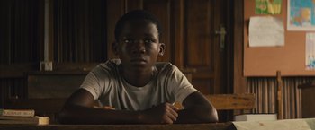 Movie still from “Beasts of No Nation” (2015), directed by Cary Joji Fukunaga – A young man sitting at a table in a dark room; Close Up shot, Low angle