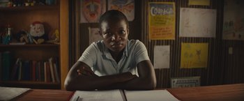 Movie still from “Beasts of No Nation” (2015), directed by Cary Joji Fukunaga – A man sitting at a table in front of papers; Medium shot, Over the shoulder angle