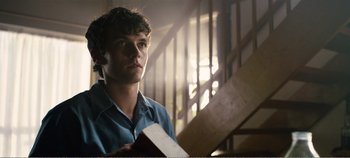 Movie still from “Black Mirror: Bandersnatch” (2018), directed by David Slade – A young man holding a book while standing next to a staircase; Close Up shot, Low angle