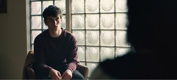 Movie still from “Black Mirror: Bandersnatch” (2018), directed by David Slade – A man sitting in front of a glass block window; Medium shot, Over the shoulder angle