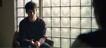 Movie still from “Black Mirror: Bandersnatch” (2018), directed by David Slade – A man sitting in front of a glass block window; Medium shot, Over the shoulder angle