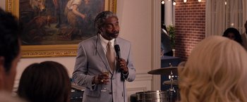 Movie still from “Bridesmaids” (2011), directed by Paul Feig – A man in a suit holding a microphone and holding a glass of wine; Medium shot, Over the shoulder angle