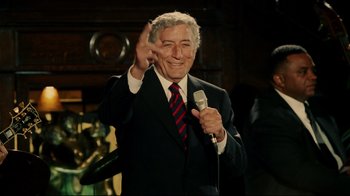 Movie still from “Bruce Almighty” (2003), directed by Tom Shadyac – A man in a suit and tie holding a microphone and waving; Medium shot, Over the shoulder angle