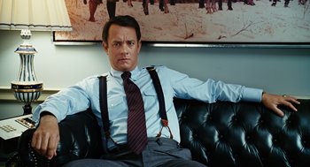 Movie still from “Charlie Wilson's War” (2007), directed by Mike Nichols – A man sitting on top of a black couch wearing suspenders; Medium shot, Over the shoulder angle
