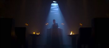 Movie still from “Chilling Adventures of Sabrina” (2018), created by Roberto Aguirre-Sacasa – A man standing at a podium in front of a stage; Wide shot, Low angle