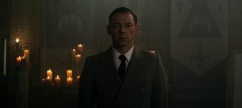 Movie still from “Chilling Adventures of Sabrina” (2018), created by Roberto Aguirre-Sacasa – A man in a suit and tie standing in front of candles in a room; Medium shot, Low angle