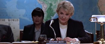 Movie still from “Clear and Present Danger” (1994), directed by Phillip Noyce – An older woman sitting in front of a microphone at a table; Close Up shot, Over the shoulder angle