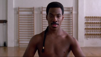 Movie still from “Coming to America” (1988), directed by John Landis – A shirtless man with a long black braid in front of a wall; Close Up shot, Over the shoulder angle