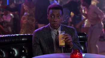 Movie still from “Coming to America” (1988), directed by John Landis – A man sitting at a table holding a glass of orange juice; Medium shot, Over the shoulder angle