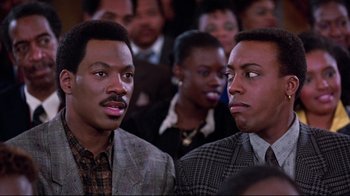 Movie still from “Coming to America” (1988), directed by John Landis – A couple of men standing next to each other in front of a crowd; Close Up shot, Over the shoulder angle